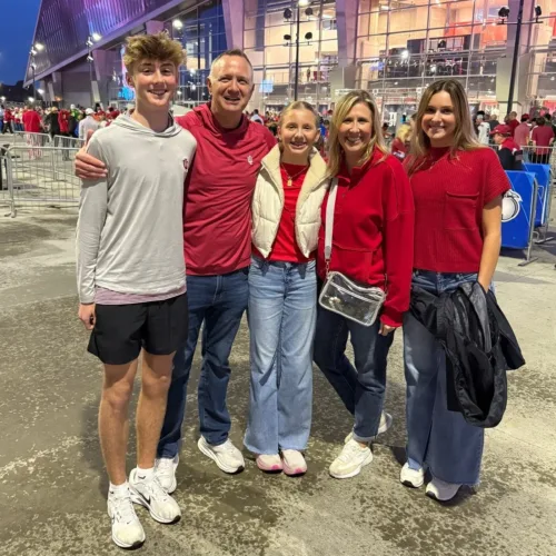A group of five people poses for a photo outside a stadium at dusk, all wearing casual clothes and red or white tops.