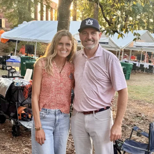 A woman and Matt Carrington stand together outdoors at a tailgate event with tents, chairs, and trees in the background.