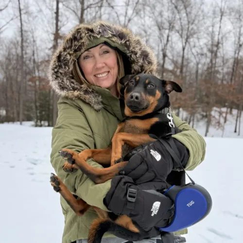 Susan Stabler, in a green winter coat, holds a medium-sized black and brown dog outside in a snowy landscape with trees in the background.