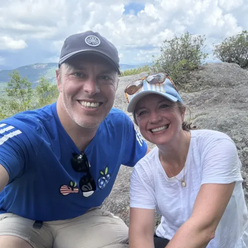 Two people smiling and posing for a selfie on a rocky mountain ledge with greenery and a cloudy sky in the background.