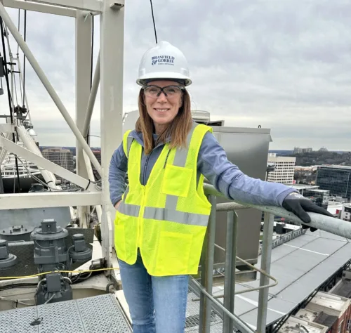 A woman wearing a hard hat, safety glasses, and a neon yellow vest stands on a metal platform at a construction site overlooking a cityscape.
