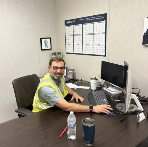 A man wearing a safety vest sits at an office desk, smiling at the camera, with a computer, water bottle, and calendar on the wall behind him.