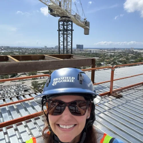 Alexia Borden, in full safety gear, takes a selfie atop a construction site rooftop with a crane and cityscape stretching out behind her.
