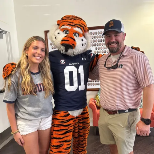 A young woman and a man stand on either side of the Auburn University tiger mascot, posing for a photo indoors.