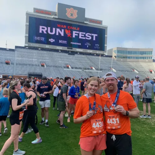 Two people wearing orange shirts and race bibs pose with medals on a football field during the War Eagle Run Fest at Auburn University.