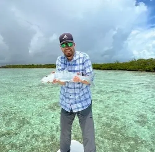 Michael Keller, wearing sunglasses and a cap, stands barefoot on a boat in clear shallow water, holding a fish with a cloudy sky and green shoreline in the background.