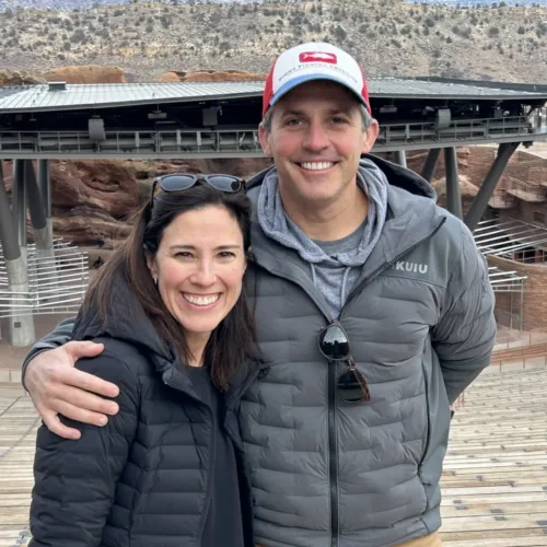 A man and woman wearing jackets and smiling stand together at an outdoor amphitheater with rocky landscape in the background.