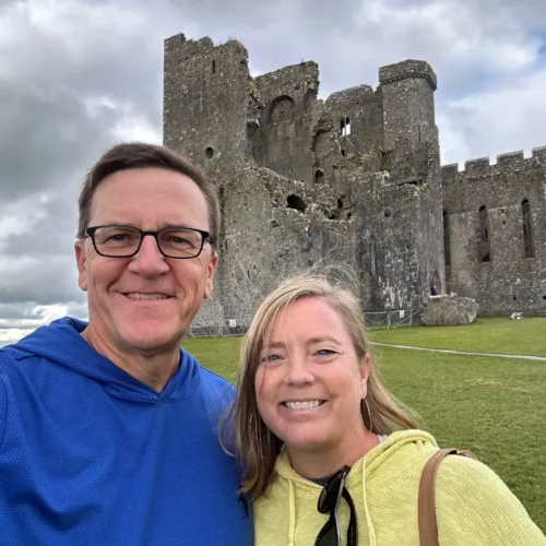 Chris Kramer and a companion smile in front of the ruins of a large stone castle on a cloudy day, with lush grass and a winding path in the foreground.