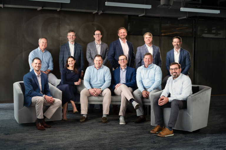 Twelve people, eleven men and one woman, pose indoors in business casual attire. Four are seated on a couch, two on chairs, and six stand behind them against a dark background.